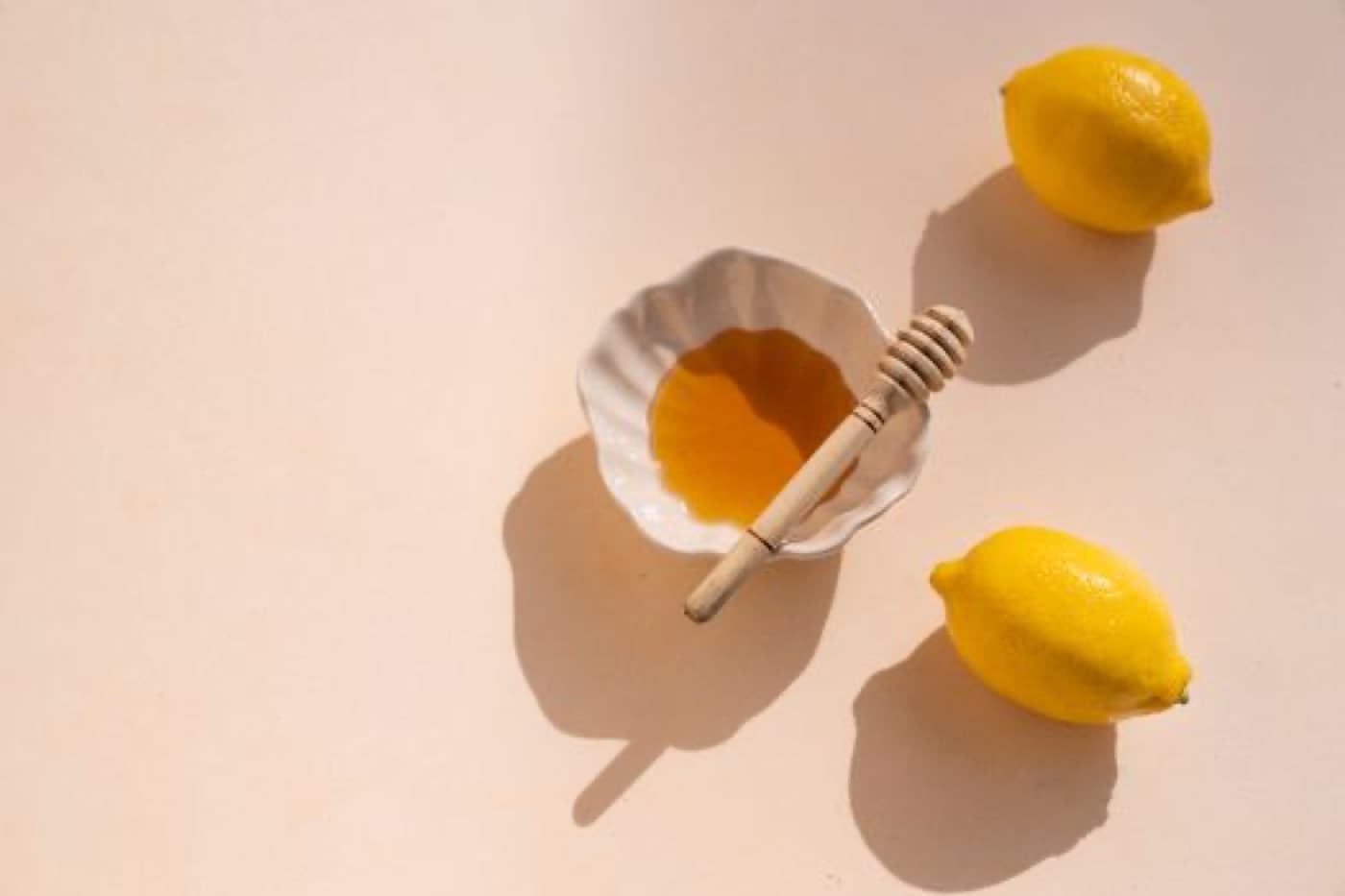 Krem rengi zeminde küçük bir kâsede bal, ahşap bal çubuğu ve iki limon