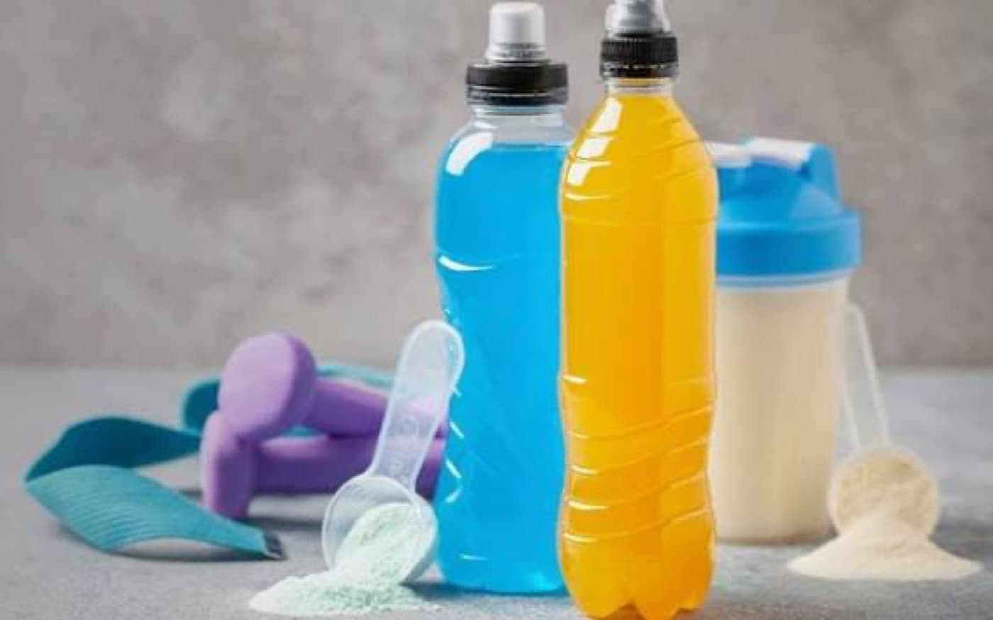 Mavi ve turuncu renkli iki spor içeceği şişesi; yanlarında dumbbell, ölçü kaşığı ve protein karıştırıcısı