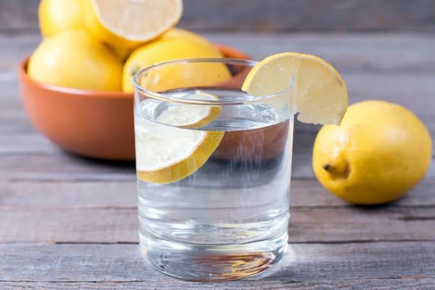 Ahşap masada, kenarına limon dilimi takılı bir bardak su ve arkada kâsede limonlar