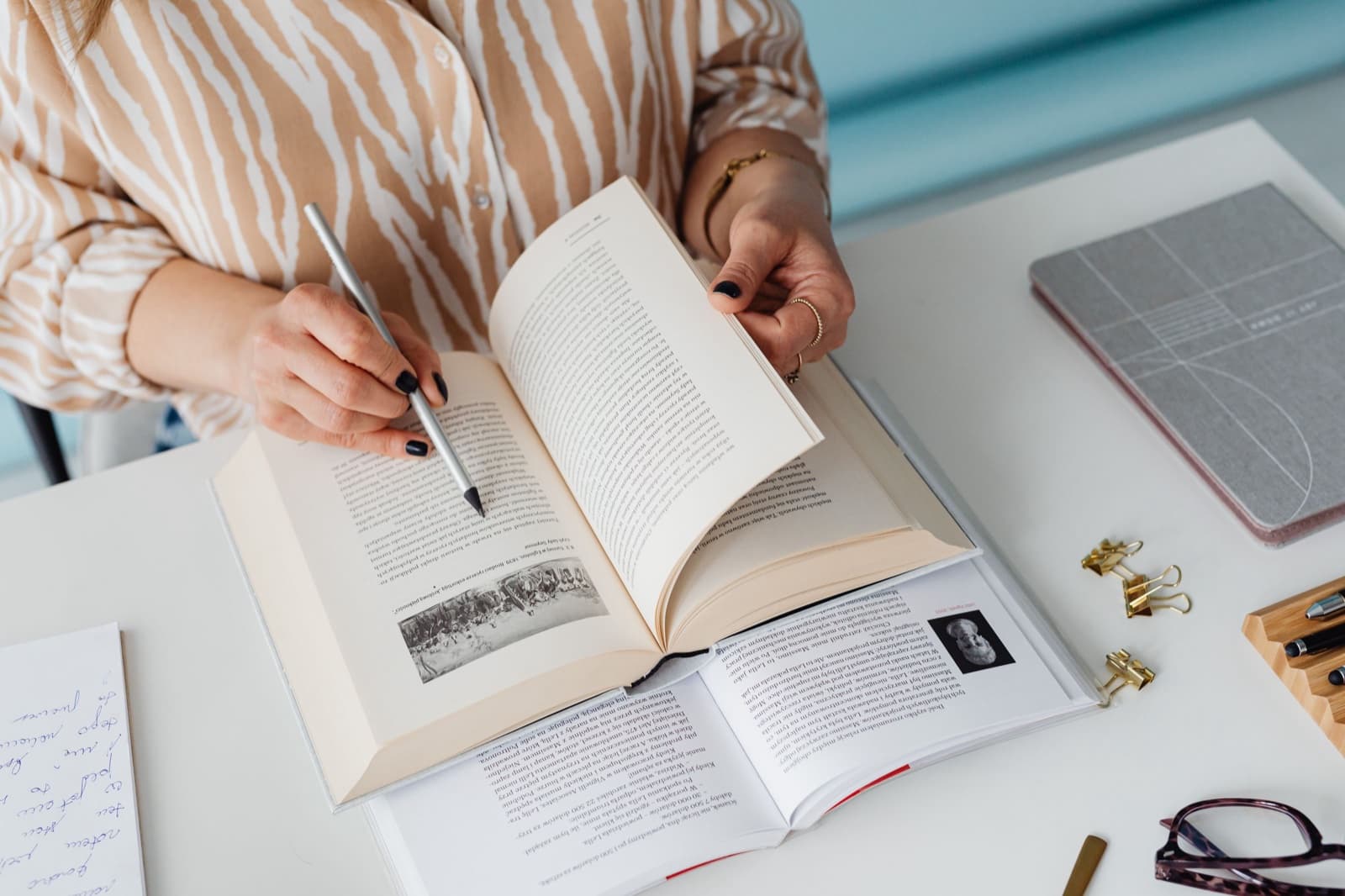 Masada elinde kalemle bir kitabı okuyan kişi; önünde ikinci bir açık kitap, el yazısı notlar ve gözlük