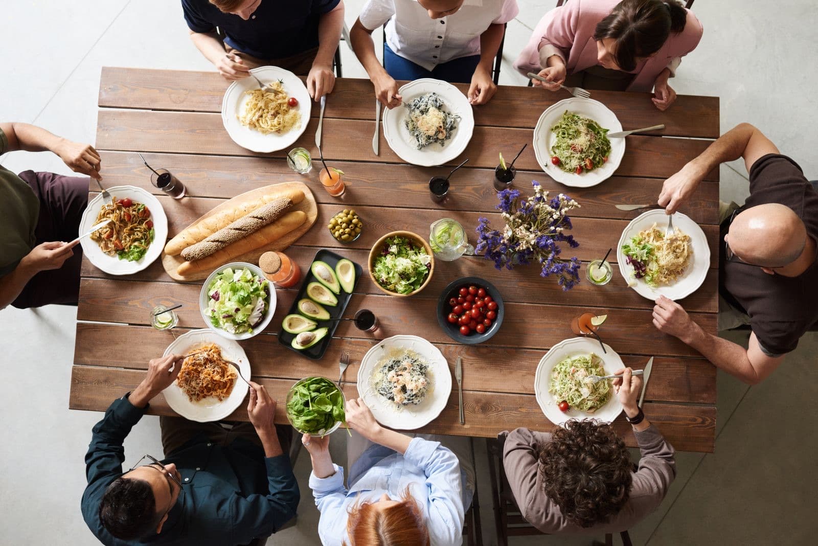 Yukarıdan görünen ahşap sofra; tabaklarda salata ve makarna, ortada uzun bir ekmek somunu, etrafında oturan kişilerin yalnızca elleri görünüyor