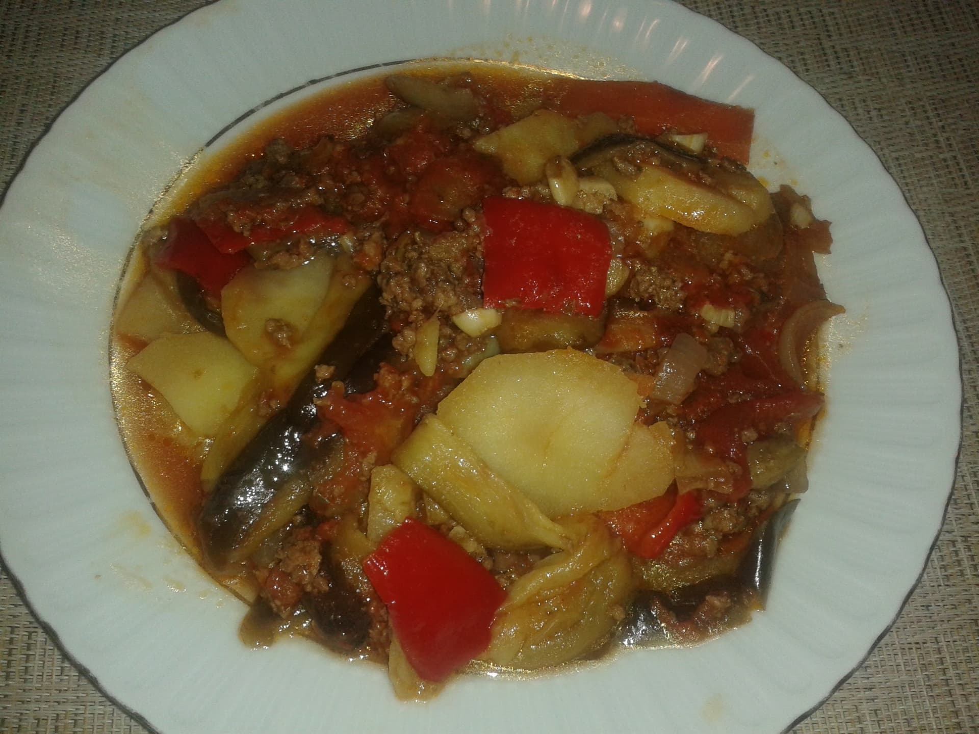 Beyaz tabakta üst üste dizilmiş patlıcan dilimleri, patates, kıyma, domates ve kırmızı biber