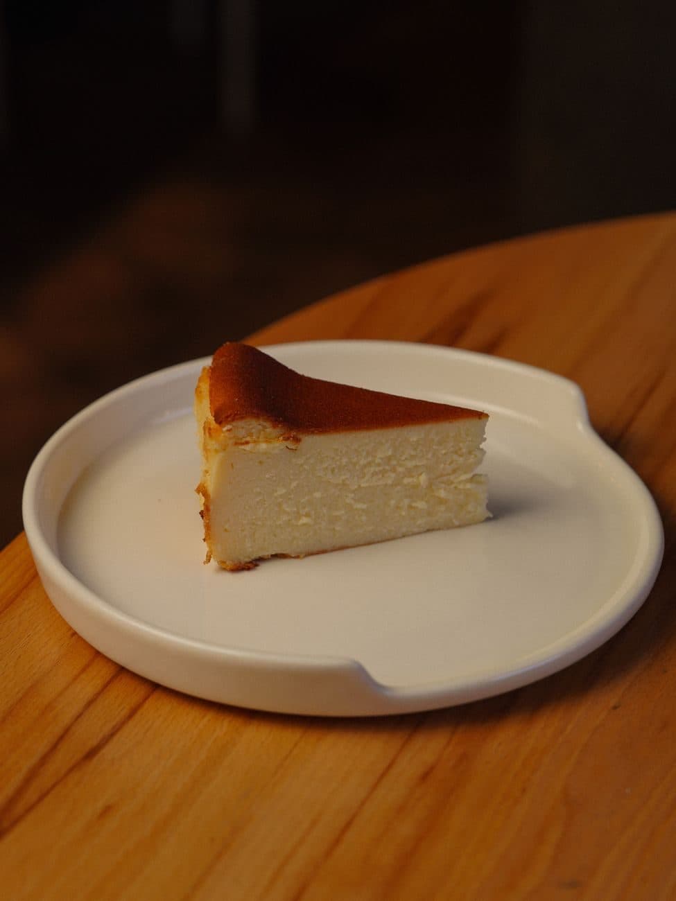 Ahşap masada beyaz tabakta, üstü sade tek dilim fırın cheesecake