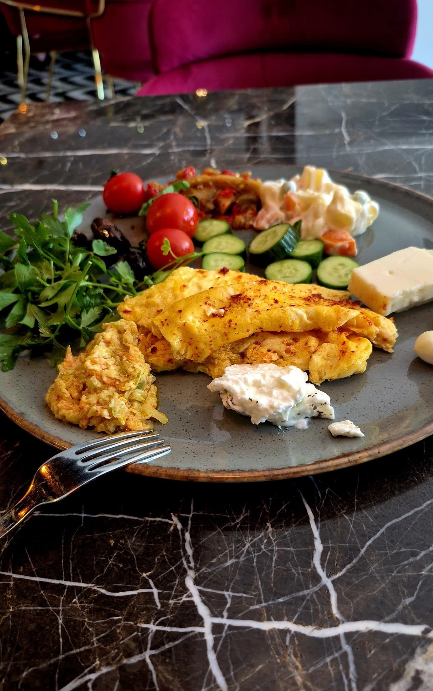 Gri tabakta katlanmış omlet, yanında beyaz peynir dilimi, yeşillik, kiraz domates ve salatalık