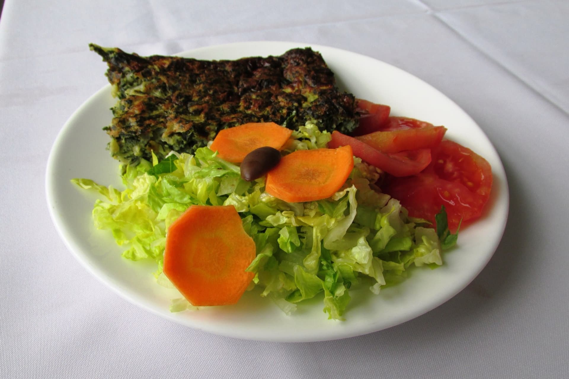 Beyaz tabakta üçgen kesilmiş koyu yeşil pazılı omlet dilimi, yanında marul, havuç ve domates