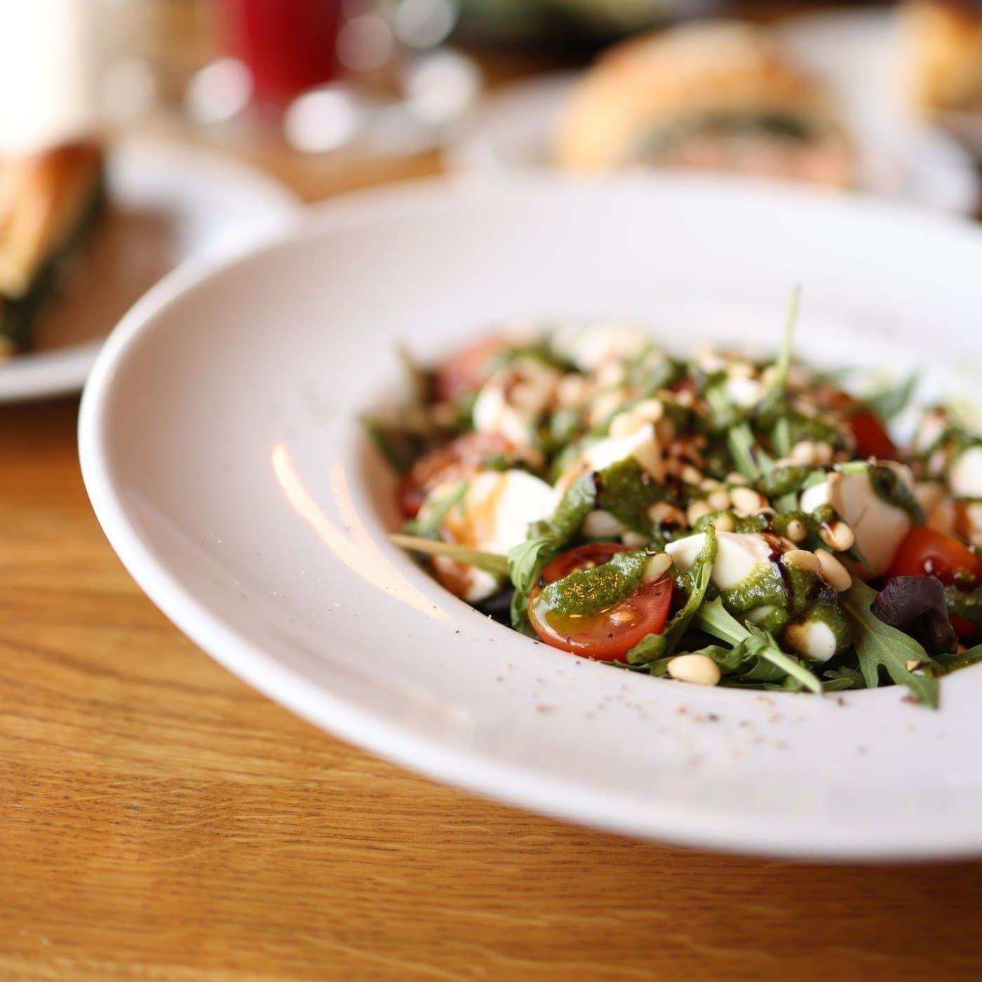 Geniş beyaz tabakta roka yaprakları ve kiraz domatesler, üzerinde mozzarella ve çam fıstığı